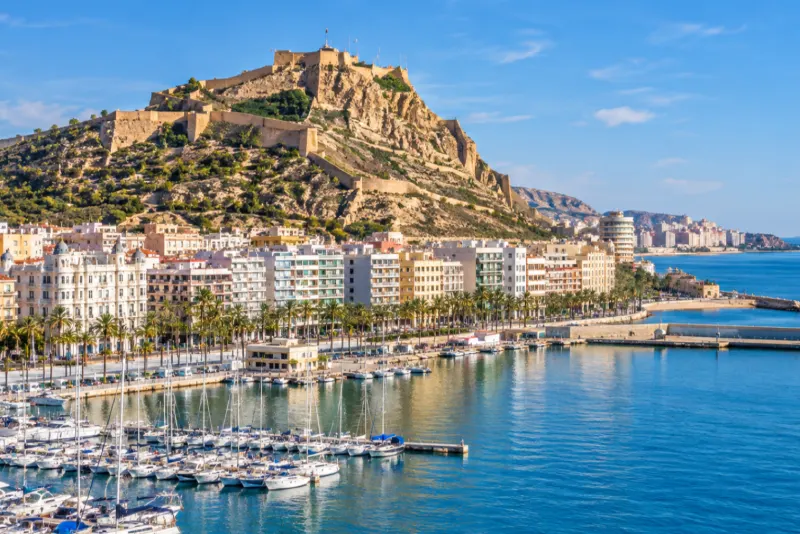 A hilltop castle above a marina and palm-lined promenade in sunshine in Alicante