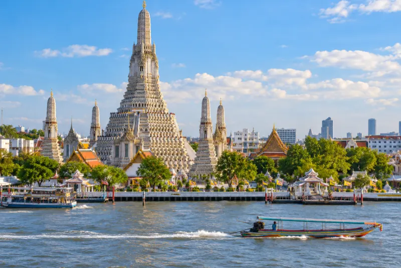 A temple with a tall carved spire beside the river and a long-tail boat in Bangkok