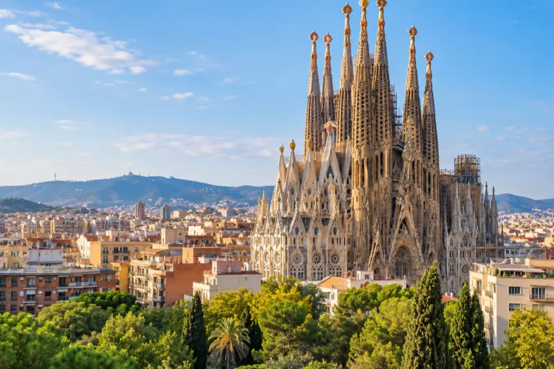 The towering spires of an unfinished basilica above rooftops and green hills in Barcelona