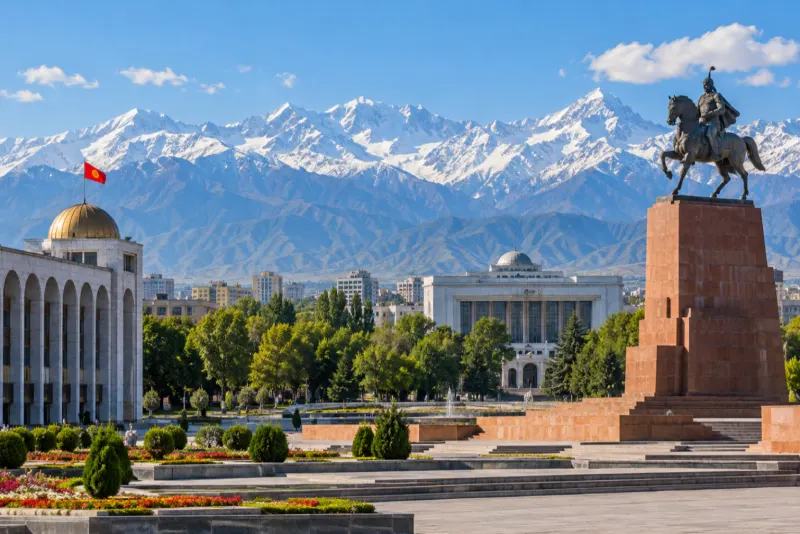A bronze horseman monument and a domed hall below a long snow-capped mountain range in Bishkek