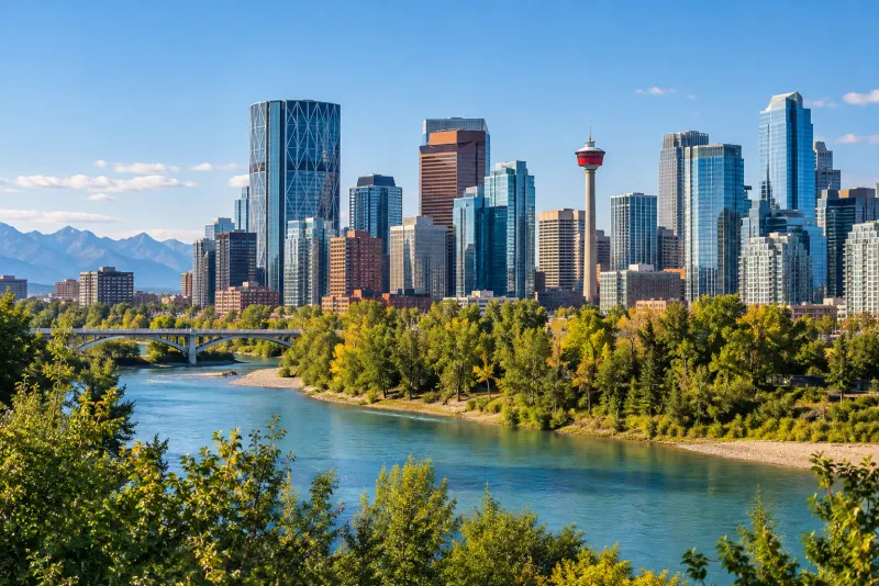 Glass towers and a red-topped tower beside a turquoise river with mountains behind in Calgary