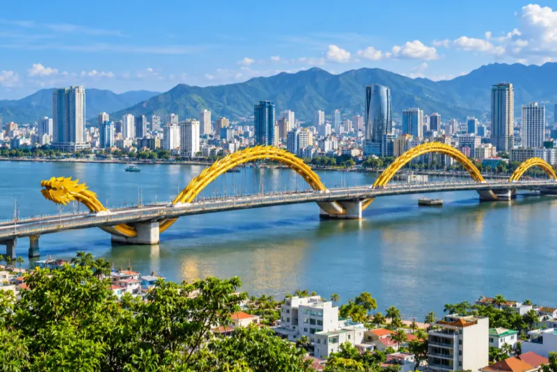 A golden dragon-shaped bridge across a river with towers and green mountains in Da Nang