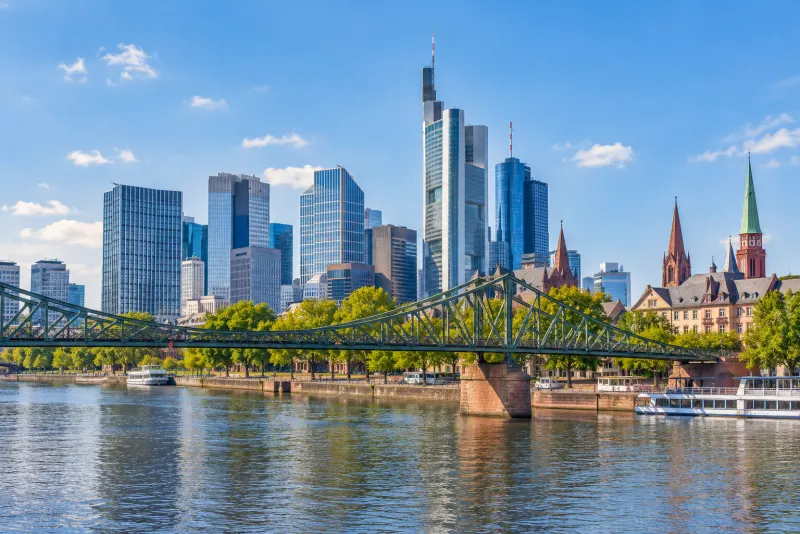 Glass skyscrapers beyond a green iron footbridge and the river in Frankfurt