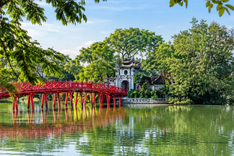 A red wooden bridge leading to a small island temple on a green lake in Hanoi