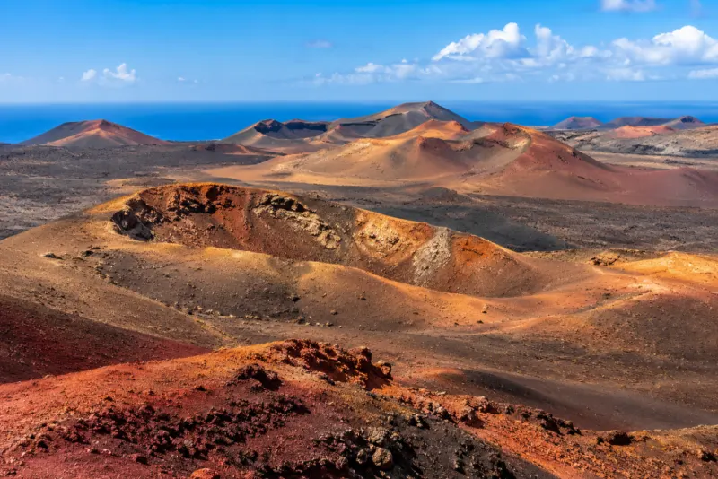 Red and orange volcanic cones and black lava fields beneath a blue sea in Lanzarote