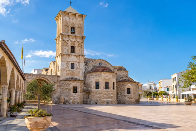 A sandstone church with a tall carved bell tower on a sunny square in Larnaca