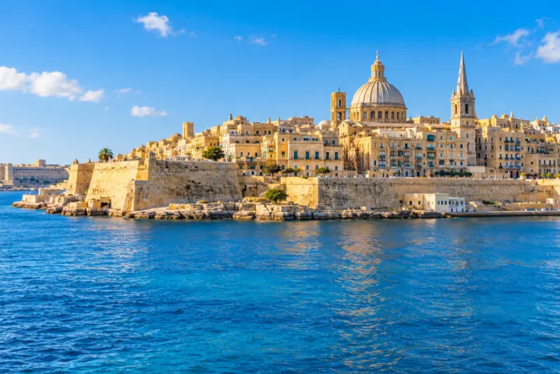 Honey-coloured fortress walls and a domed church above blue harbour water in Malta
