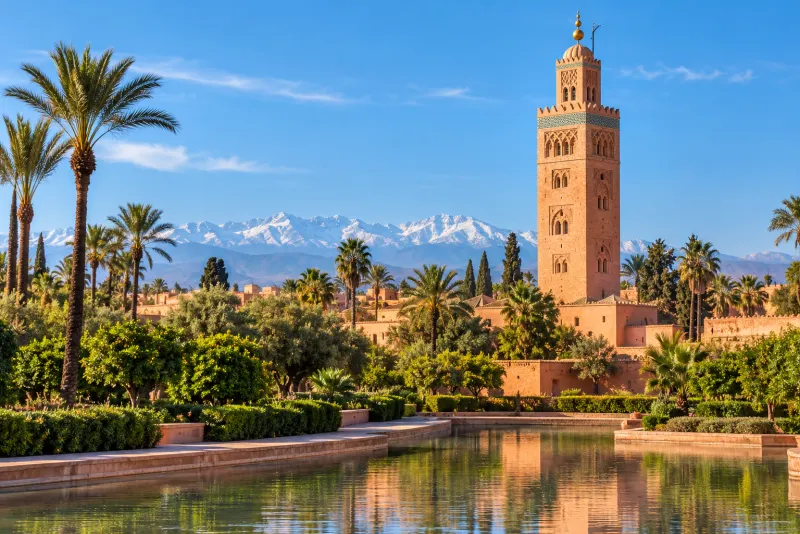 A tall red sandstone minaret and palms by a reflecting pool before snowy mountains in Marrakech