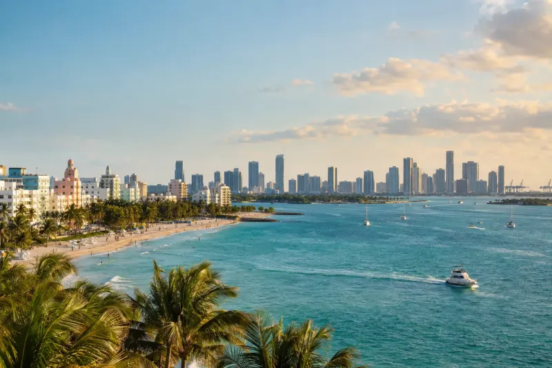 A palm-fringed beach with pastel art deco buildings and a skyline across the bay in Miami