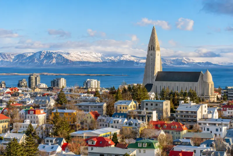 A tall modern church above colourful rooftops with snowy mountains across the bay in Reykjavik