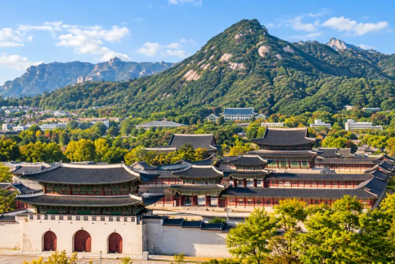 A traditional palace with tiered tiled roofs below a forested granite mountain in Seoul