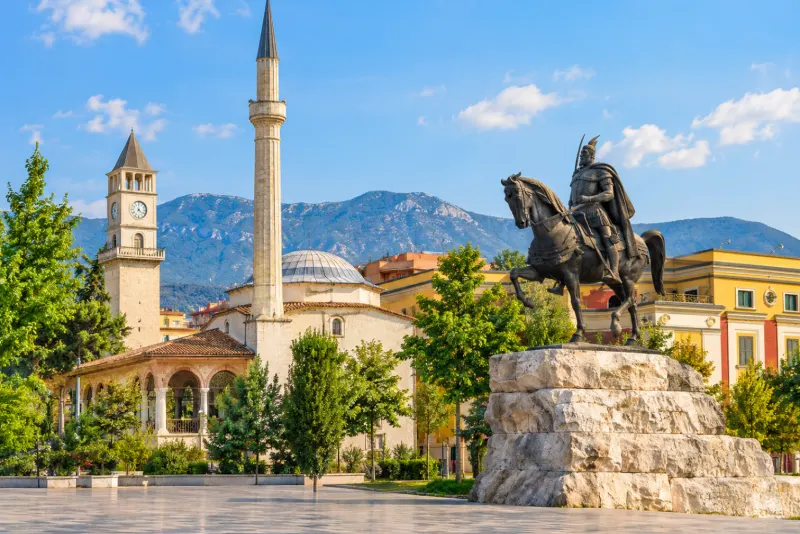 A bronze horseman statue, a clock tower and a minaret on a sunny square in Tirana