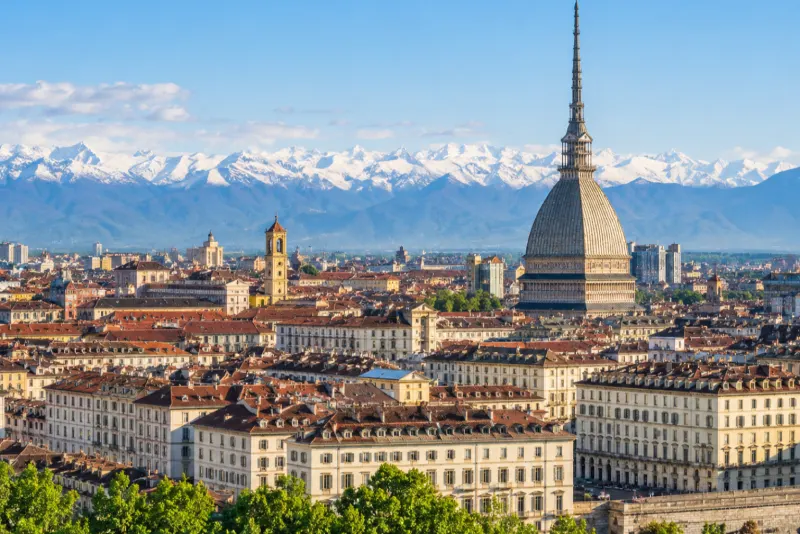 A tall spired dome rising above red rooftops with snowy Alps beyond in Turin