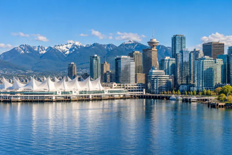 White sail-roofed waterfront terminal and glass towers below snowy mountains in Vancouver